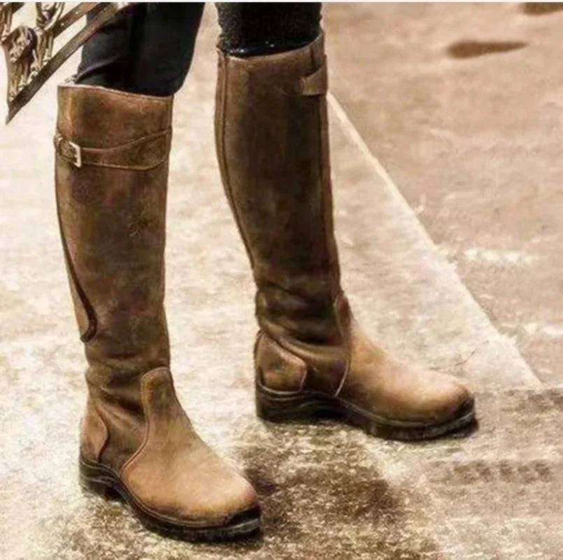 Une paire de bottes en cuir marron, hauteur genou, pour femmes, avec un détail de boucle sur le côté et une semelle robuste, parfaites pour l'hiver et les activités en plein air.